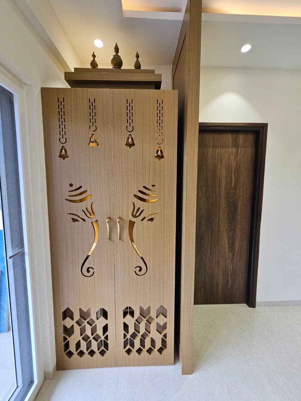 Pooja unit with a cut-out Ganesha motif and brass bells in an apartment interior by ACIPL
